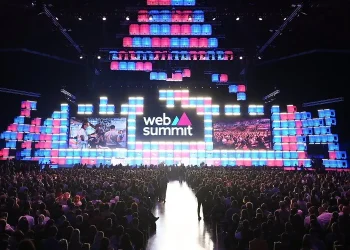La grande folla di fronte al palco del Forum di Parque das Nações il durante il Web Summit di Lisbona 2024. Lo sfondo è caratterizzato da pannelli luminosi rettangolari colorati in tonalità blu, rosa e viola. Due grandi schermi mostrano le immagini dell'evento.