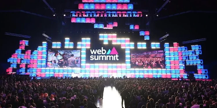La grande folla di fronte al palco del Forum di Parque das Nações il durante il Web Summit di Lisbona 2024. Lo sfondo è caratterizzato da pannelli luminosi rettangolari colorati in tonalità blu, rosa e viola. Due grandi schermi mostrano le immagini dell'evento.