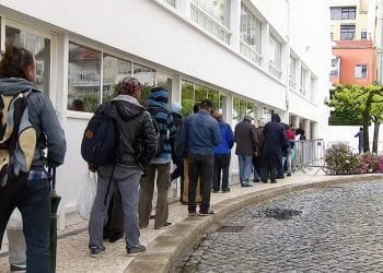 Le persone stanno in fila indiana accanto a un edificio con grandi finestre. La maggior parte indossa giacche e zaini. Il marciapiede curva e ci sono fiori e pozzanghere sulla strada acciottolata. Sullo sfondo sono visibili edifici a più piani e alberi del Portogallo, che ne suggeriscono la bellezza in mezzo alla povertà.