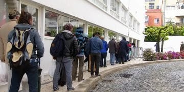 Le persone stanno in fila indiana accanto a un edificio con grandi finestre. La maggior parte indossa giacche e zaini. Il marciapiede curva e ci sono fiori e pozzanghere sulla strada acciottolata. Sullo sfondo sono visibili edifici a più piani e alberi del Portogallo, che ne suggeriscono la bellezza in mezzo alla povertà.