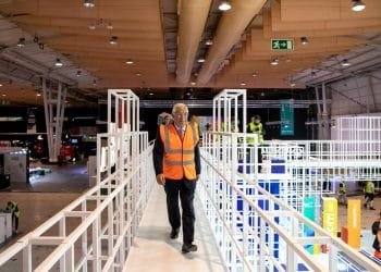 Un uomo in abito e gilet ad alta visibilità cammina su una piattaforma di metallo bianco all'interno di un grande centro congressi, con molte altre persone con gilet simili che lavorano sullo sfondo. Il soffitto è rivestito con luci sospese e condotti di ventilazione, una scena che ricorda il vivace evento dei Quattro Giorni Lavorativi in Portogallo.