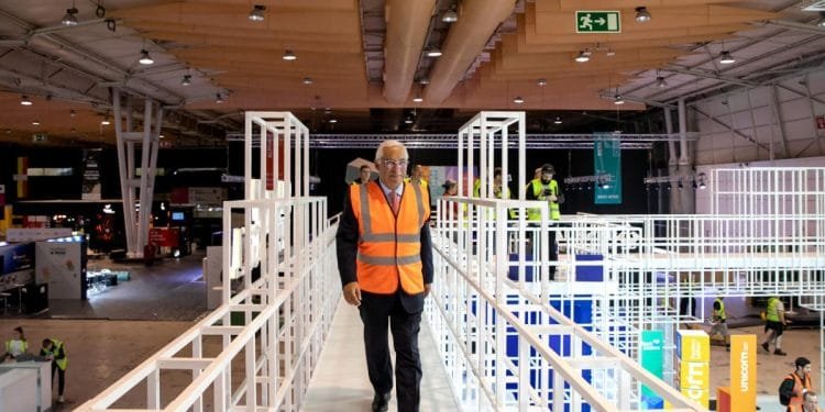 Un uomo in abito e gilet ad alta visibilità cammina su una piattaforma di metallo bianco all'interno di un grande centro congressi, con molte altre persone con gilet simili che lavorano sullo sfondo. Il soffitto è rivestito con luci sospese e condotti di ventilazione, una scena che ricorda il vivace evento dei Quattro Giorni Lavorativi in Portogallo.
