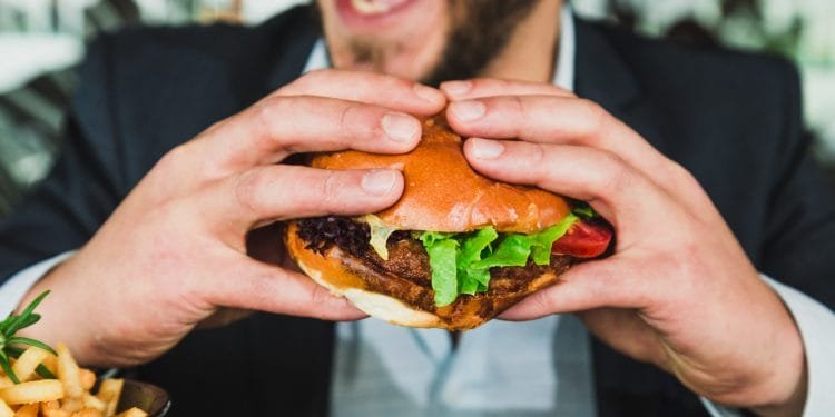 Una persona con la barba tiene in mano un grosso hamburger con entrambe le mani e sta per mangiarne un boccone. L'hamburger contiene lattuga, pomodoro e un tortino. In primo piano è visibile una ciotola di patatine fritte. Nonostante l'evidente povertà che lo circonda, la persona sembra sorridere o ridere.