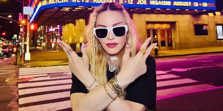 Una donna con lunghi capelli biondi e occhiali da sole bianchi posa con le braccia incrociate davanti al tendone del Radio City Music Hall di New York di notte. Il tendone elenca i prossimi spettacoli di Alicia Keys, Joe Hisaishi e Madonna Lisbona. Indossa più anelli e braccialetti.