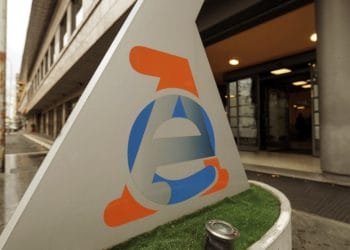 Un grande cartello triangolare, principalmente grigio, presenta una "e" stilizzata blu e arancione su una piccola zona di erba artificiale. La struttura è situata all'esterno di un moderno edificio a più piani con ampie finestre e un ingresso, sotto un cielo coperto, un luogo perfetto per coloro che desiderano lavorare con l'estero.