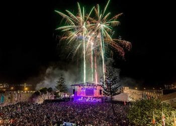 Un vibrante spettacolo pirotecnico illumina il cielo notturno sopra un grande concerto all'aperto al Festival Músicas do Mundo. Il palco, inondato di luci colorate, è circondato da una folla enorme di spettatori. Alberi e bandiere sono visibili in primo piano, contribuendo a creare un'atmosfera festosa.