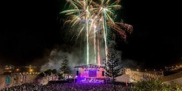 Un vibrante spettacolo pirotecnico illumina il cielo notturno sopra un grande concerto all'aperto al Festival Músicas do Mundo. Il palco, inondato di luci colorate, è circondato da una folla enorme di spettatori. Alberi e bandiere sono visibili in primo piano, contribuendo a creare un'atmosfera festosa.