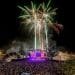Un vibrante spettacolo pirotecnico illumina il cielo notturno sopra un grande concerto all'aperto al Festival Músicas do Mundo. Il palco, inondato di luci colorate, è circondato da una folla enorme di spettatori. Alberi e bandiere sono visibili in primo piano, contribuendo a creare un'atmosfera festosa.