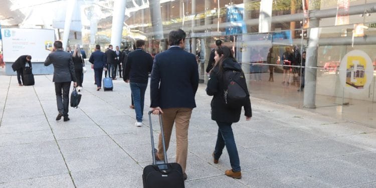 La gente cammina lungo un percorso all'aperto accanto a un moderno edificio in vetro alla Fiera Turismo Lisbona. Alcune persone trascinano le valigie. La scena appare affollata, con persone vestite con un mix di abiti da lavoro e casual. L'area è luminosa, suggerendo che sia giorno.