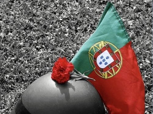 L'elmetto di un soldato poggia sull'erba accanto a un garofano rosso e una bandiera portoghese. Il fiore e la bandiera risaltano con colori vivaci sullo sfondo bianco e nero, a simboleggiare la rivoluzione dei garofani in Portogallo. Questa immagine toccante fa parte di una mostra fotografica dell'Algarve, che cattura momenti di significato storico.