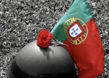 L'elmetto di un soldato poggia sull'erba accanto a un garofano rosso e una bandiera portoghese. Il fiore e la bandiera risaltano con colori vivaci sullo sfondo bianco e nero, a simboleggiare la rivoluzione dei garofani in Portogallo. Questa immagine toccante fa parte di una mostra fotografica dell'Algarve, che cattura momenti di significato storico.