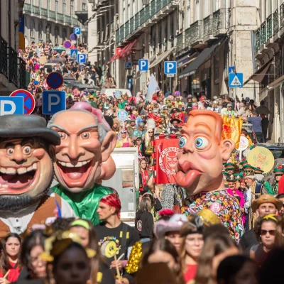 Una vivace scena di strada cattura l'essenza del carnevale di Torres Vedras in Portogallo, con una grande folla che guarda la sfilata colorata. Le persone si radunano sui marciapiedi vicino a un edificio mentre palloncini e costumi vivaci riempiono l'aria. Una figura gigante e una decorazione blu aleggiano sopra, aggiungendo spirito festoso.