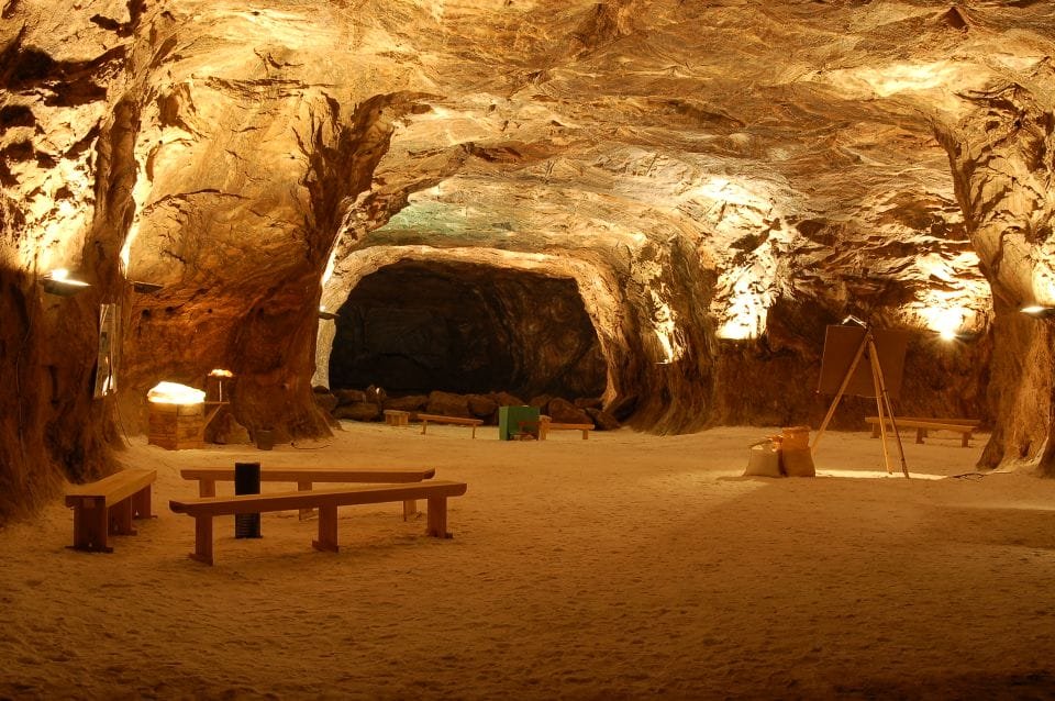 Una grotta sotterranea spaziosa e ben illuminata, con pareti rocciose strutturate che ricordano le affascinanti "attrazioni dell'Algarve". L'area è caratterizzata da diverse panche di legno, una piccola lavagna su cavalletto e vari oggetti sparsi qua e là, il tutto posizionato su un pavimento sabbioso. L'illuminazione calda crea un'atmosfera accogliente.