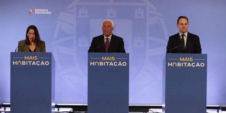 Tre persone stanno dietro i podi durante una conferenza stampa. I podi hanno la scritta "MAIS HABITAÇÃO" e lo sfondo include il logo e il testo "REPÚBLICA PORTUGUESA". Le persone sembrano rivolgersi al pubblico riguardo alle misure abitazione.
