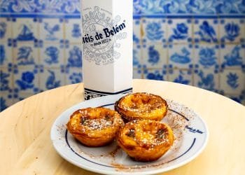 Un piatto di tre Pastéis de Belém, tradizionali crostate portoghesi con crema pasticcera, è seduto su un tavolo di legno chiaro. Sullo sfondo c'è una scatola bianca con il logo "Pastel de Belém". Lo sfondo è decorato con piastrelle di ceramica portoghese blu e bianche.