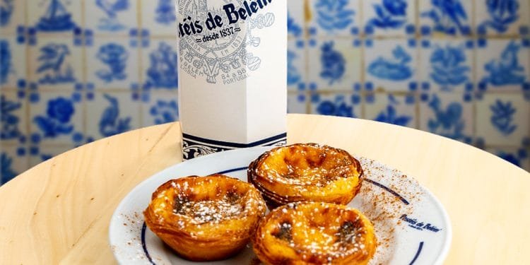 Un piatto di tre Pastéis de Belém, tradizionali crostate portoghesi con crema pasticcera, è seduto su un tavolo di legno chiaro. Sullo sfondo c'è una scatola bianca con il logo "Pastel de Belém". Lo sfondo è decorato con piastrelle di ceramica portoghese blu e bianche.
