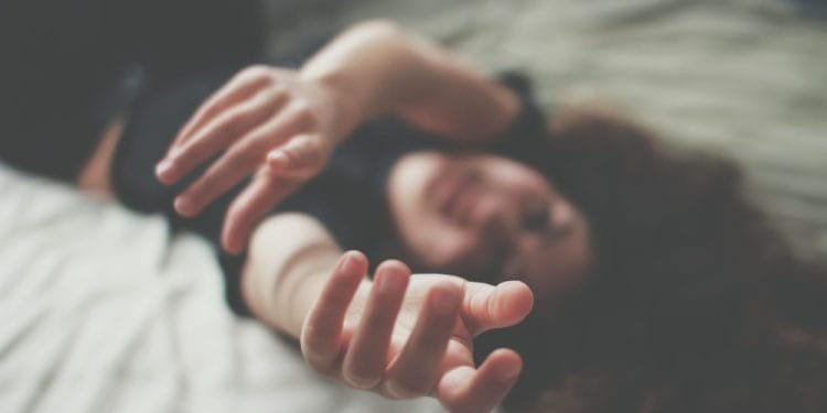 Una persona giace supina su un letto con le mani protese verso la fotocamera in un primo piano sfocato. Lo sfondo è sfocato, enfatizzando le mani e creando un'atmosfera sognante o rilassata, lontana dalla dura realtà come la violenza domestica in Portogallo. La persona ha i capelli lunghi e ricci.