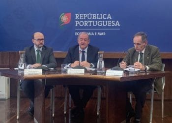 Tre uomini sono seduti al tavolo di una sala conferenze. L'uomo al centro parla al microfono, mentre gli altri due ascoltano attentamente. Sul fondale sono presenti il logo e il nome della "República Portuguesa" e "Economia e Mar", oltre ai riferimenti all'ASAE controlli prezzi scritti in portoghese.