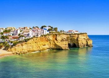 Una vista panoramica sulla costa che mostra una cittadina sulla scogliera con edifici imbiancati nell'Algarve. Le scogliere si affacciano su un mare calmo e turchese con alla base una spiaggia sabbiosa, rendendolo perfetto per una visita a febbraio. Il cielo azzurro e limpido esalta i colori vivaci del paesaggio.