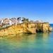 Una vista panoramica sulla costa che mostra una cittadina sulla scogliera con edifici imbiancati nell'Algarve. Le scogliere si affacciano su un mare calmo e turchese con alla base una spiaggia sabbiosa, rendendolo perfetto per una visita a febbraio. Il cielo azzurro e limpido esalta i colori vivaci del paesaggio.