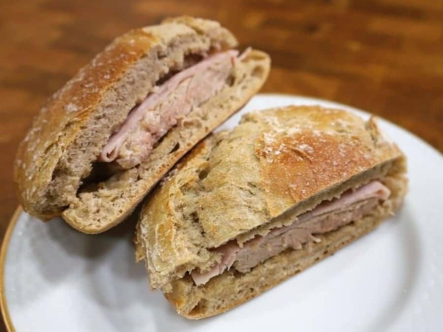 Un panino Bifana con fette di carne, possibilmente prosciutto, tra metà di un panino integrale croccante, servito su un piatto bianco con una superficie di tavolo in legno sullo sfondo.