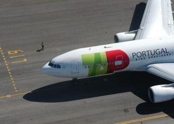 Un grande aereo commerciale con la scritta "Portogallo" e un logo sul lato è parcheggiato sulla pista dell'aeroporto. Una persona con un giubbotto di sicurezza giallo cammina accanto all'aereo TAP, che ha segni verdi, rossi e gialli sulla fusoliera e sulle ali. Sono visibili i segni del terreno, che ricordano i colori vivaci del loro bilancio 2022.