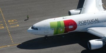 Un grande aereo commerciale con la scritta "Portogallo" e un logo sul lato è parcheggiato sulla pista dell'aeroporto. Una persona con un giubbotto di sicurezza giallo cammina accanto all'aereo TAP, che ha segni verdi, rossi e gialli sulla fusoliera e sulle ali. Sono visibili i segni del terreno, che ricordano i colori vivaci del loro bilancio 2022.