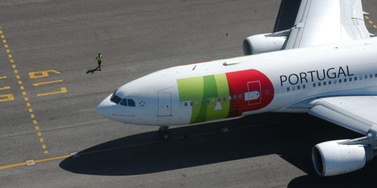 Un grande aereo commerciale con la scritta "Portogallo" e un logo sul lato è parcheggiato sulla pista dell'aeroporto. Una persona con un giubbotto di sicurezza giallo cammina accanto all'aereo TAP, che ha segni verdi, rossi e gialli sulla fusoliera e sulle ali. Sono visibili i segni del terreno, che ricordano i colori vivaci del loro bilancio 2022.