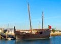 Una replica dello storico veliero in legno Caravela Boa Esperança è ormeggiata in un porto turistico sotto un cielo azzurro. La nave è dotata di alti alberi, vele e bandiera portoghese. Sullo sfondo si vedono barche a vela e altre imbarcazioni, insieme al frangiflutti in pietra del porto turistico.