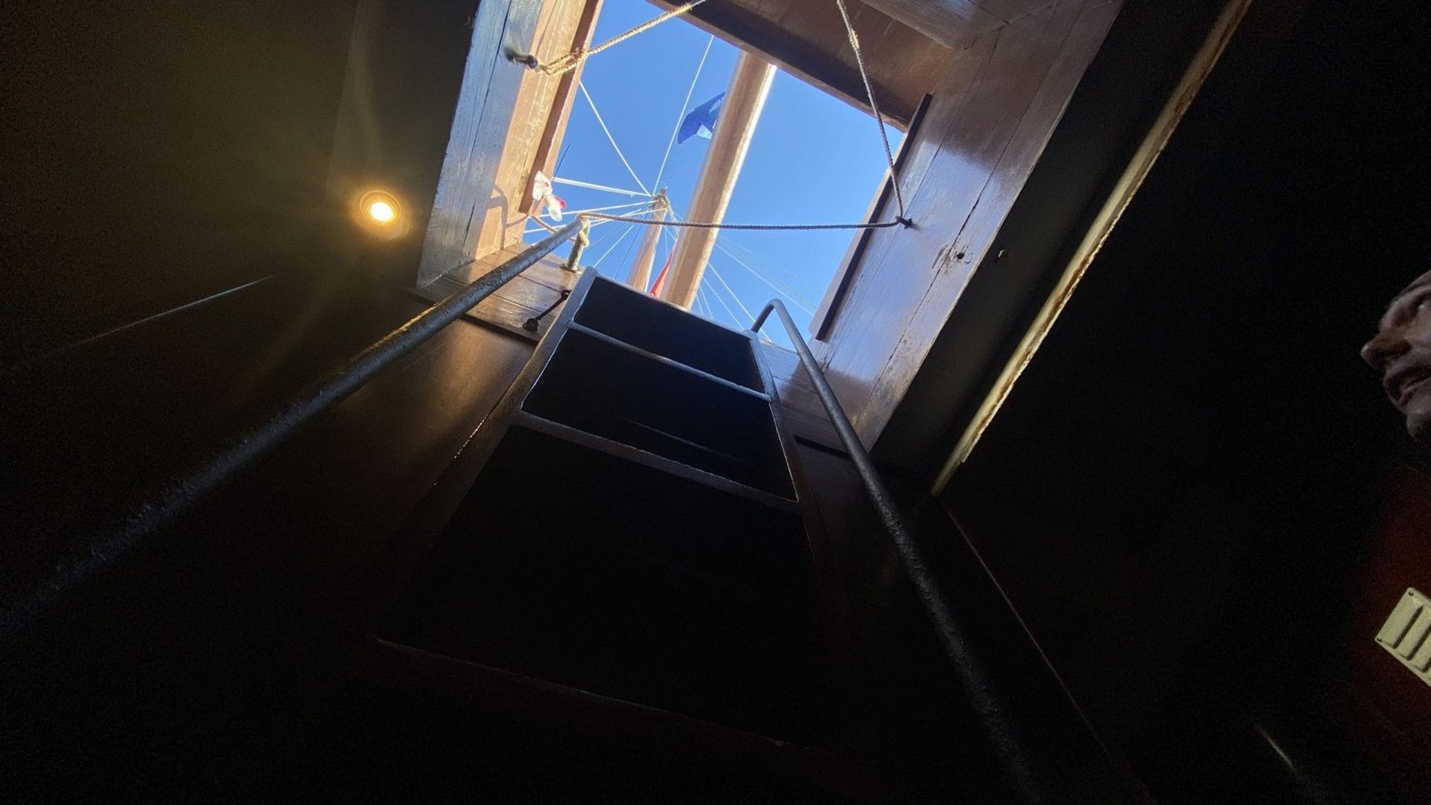 Una scala ripida che conduce ad un portello aperto rivela uno scorcio dell'esterno. Il cielo è parzialmente visibile, insieme a parte di una bandiera della Caravela Boa Esperança Centro Interpretativo. L'illuminazione all'interno è fioca, in contrasto con la luce del giorno all'esterno. Una persona è parzialmente visibile sul lato destro dell'immagine.