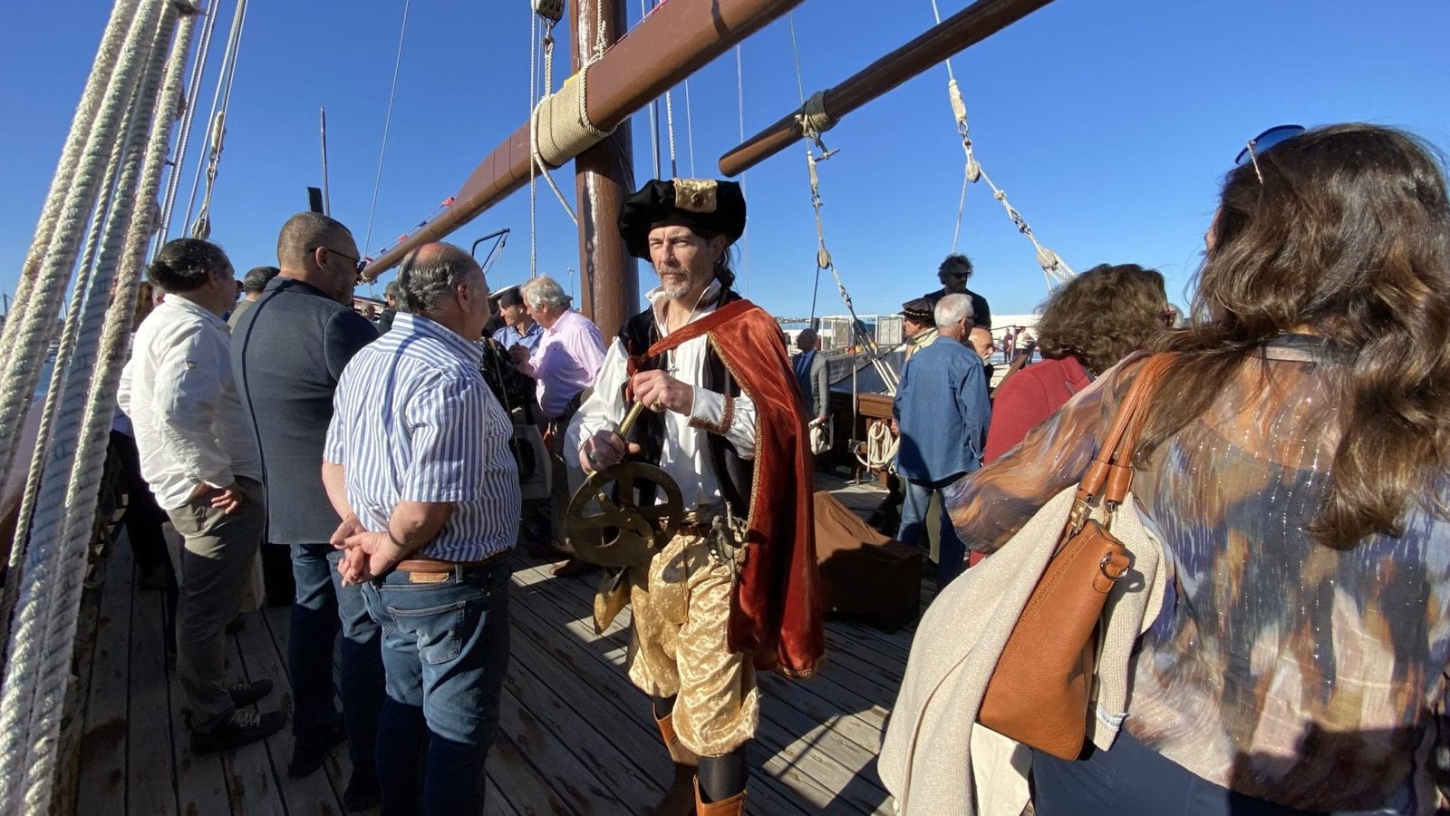 Un uomo vestito da pirata o navigatore storico, con in mano uno strumento di navigazione, si trova in mezzo a un gruppo di persone sul ponte della Caravela Boa Esperança. Il gruppo sembra interagire e godersi l'esperienza sotto un cielo azzurro e limpido al Centro Interpretativo.