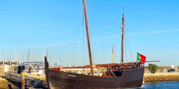 Una replica dello storico veliero in legno Caravela Boa Esperança è ormeggiata in un porto turistico sotto un cielo azzurro. La nave è dotata di alti alberi, vele e bandiera portoghese. Sullo sfondo si vedono barche a vela e altre imbarcazioni, insieme al frangiflutti in pietra del porto turistico.