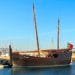 Una replica dello storico veliero in legno Caravela Boa Esperança è ormeggiata in un porto turistico sotto un cielo azzurro. La nave è dotata di alti alberi, vele e bandiera portoghese. Sullo sfondo si vedono barche a vela e altre imbarcazioni, insieme al frangiflutti in pietra del porto turistico.