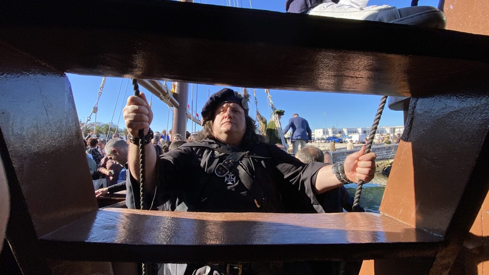Una persona vestita con abiti storici, tra cui un mantello e un cappello neri, si trova sul ponte di legno della Caravela Boa Esperança Centro Interpretativo. Tengono delle corde e sembrano seri, mentre altre persone e gli edifici del porto sono visibili sullo sfondo.