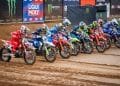 Un gruppo di piloti di motocross in abiti e caschi colorati si schiera al cancello di partenza, pronto per iniziare una gara di motociclismo di Coppa Mondo. La strada sterrata è bagnata e fangosa e sullo sfondo sono visibili vari striscioni di marchi, tra cui Liqui Moly e Monster Energy.