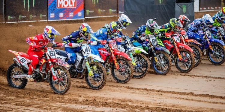 Un gruppo di piloti di motocross in abiti e caschi colorati si schiera al cancello di partenza, pronto per iniziare una gara di motociclismo di Coppa Mondo. La strada sterrata è bagnata e fangosa e sullo sfondo sono visibili vari striscioni di marchi, tra cui Liqui Moly e Monster Energy.