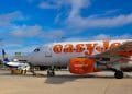 Un aereo easyJet parcheggiato si trova sulla pista di un aeroporto sotto un cielo parzialmente nuvoloso. Il personale di terra e i veicoli sono visibili vicino all'aereo, che fa parte della base EasyJet. Sullo sfondo si può vedere un aereo Ryanair tra le strutture aeroportuali e la segnaletica della proprietà di Faro.
