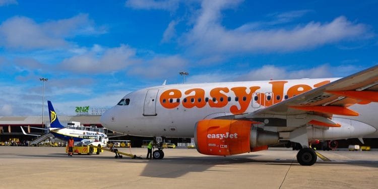 Un aereo easyJet parcheggiato si trova sulla pista di un aeroporto sotto un cielo parzialmente nuvoloso. Il personale di terra e i veicoli sono visibili vicino all'aereo, che fa parte della base EasyJet. Sullo sfondo si può vedere un aereo Ryanair tra le strutture aeroportuali e la segnaletica della proprietà di Faro.