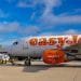 Un aereo easyJet parcheggiato si trova sulla pista di un aeroporto sotto un cielo parzialmente nuvoloso. Il personale di terra e i veicoli sono visibili vicino all'aereo, che fa parte della base EasyJet. Sullo sfondo si può vedere un aereo Ryanair tra le strutture aeroportuali e la segnaletica della proprietà di Faro.