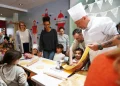 Chef Luca in uniforme bianca mostra come fare la pasta con una macchina per la pasta a bambini e adulti riuniti intorno a un tavolo all' Evento Comites Portogallo di Faro, in una sala decorata con colori vivaci.