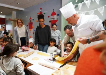 Chef Luca in uniforme bianca mostra come fare la pasta con una macchina per la pasta a bambini e adulti riuniti intorno a un tavolo all' Evento Comites Portogallo di Faro, in una sala decorata con colori vivaci.