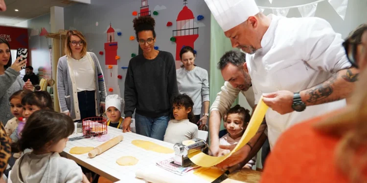Chef Luca in uniforme bianca mostra come fare la pasta con una macchina per la pasta a bambini e adulti riuniti intorno a un tavolo all' Evento Comites Portogallo di Faro, in una sala decorata con colori vivaci.