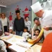 Chef Luca in uniforme bianca mostra come fare la pasta con una macchina per la pasta a bambini e adulti riuniti intorno a un tavolo all' Evento Comites Portogallo di Faro, in una sala decorata con colori vivaci.