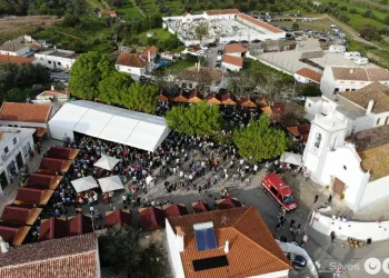 Vista aerea di un affollato del Festival del Folar dolce pasquale portoghese a Silves. Immagine all'aperto con tende bianche e tettoie arancioni tra edifici bianchi, tra cui una chiesa, dove i visitatori possono gustare prelibatezze come il Folar, circondati dal verde e dai veicoli parcheggiati.