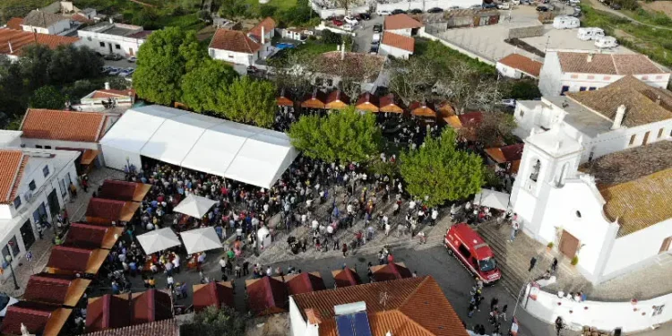 Vista aerea di un affollato del Festival del Folar dolce pasquale portoghese a Silves. Immagine all'aperto con tende bianche e tettoie arancioni tra edifici bianchi, tra cui una chiesa, dove i visitatori possono gustare prelibatezze come il Folar, circondati dal verde e dai veicoli parcheggiati.