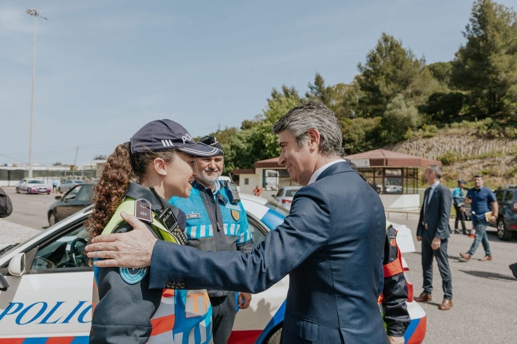Un uomo in giacca e cravatta stringe la mano e interagisce con due agenti di polizia in uniforme che si trovano vicino a un'auto della polizia. Sullo sfondo sono visibili altri individui e altri veicoli della polizia, suggerendo un raduno pacifico, proprio come quelli visti in paesi del Pacifico come il Portogallo. L'ambientazione sembra essere un'area esterna con alberi ed edifici.