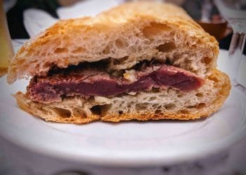 Immagine del primo piano di un panino con bistecca su un piatto bianco. Il panino presenta una spessa fetta di bistecca al sangue incastonata tra due metà di una baguette croccante, che ricorda una bifana portoghese, mettendo in mostra la carne rosa e succosa e la consistenza ariosa del pane.