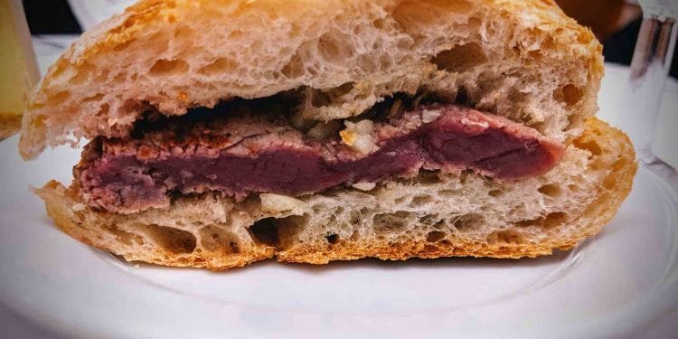 Immagine del primo piano di un panino con bistecca su un piatto bianco. Il panino presenta una spessa fetta di bistecca al sangue incastonata tra due metà di una baguette croccante, che ricorda una bifana portoghese, mettendo in mostra la carne rosa e succosa e la consistenza ariosa del pane.