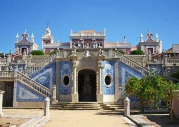 Un grande edificio storico con un'architettura ricercata e accenti di piastrelle blu è catturato sotto un cielo azzurro e limpido. Ampie scale in pietra con intricate ringhiere conducono ad un ingresso ad arco fiancheggiato da due finestre circolari. Una vegetazione lussureggiante circonda la scena, che ricorda il fascino che si prova quando si visitano gli aldeias dell'Algarve.