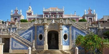 Un grande edificio storico con un'architettura ricercata e accenti di piastrelle blu è catturato sotto un cielo azzurro e limpido. Ampie scale in pietra con intricate ringhiere conducono ad un ingresso ad arco fiancheggiato da due finestre circolari. Una vegetazione lussureggiante circonda la scena, che ricorda il fascino che si prova quando si visitano gli aldeias dell'Algarve.